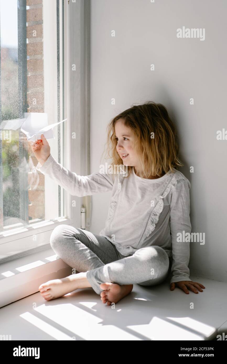 Felice bambina bionda in pigiama giocando con aereo di carta mentre si siede a piedi nudi sul davanzale della finestra il giorno di sole Foto Stock