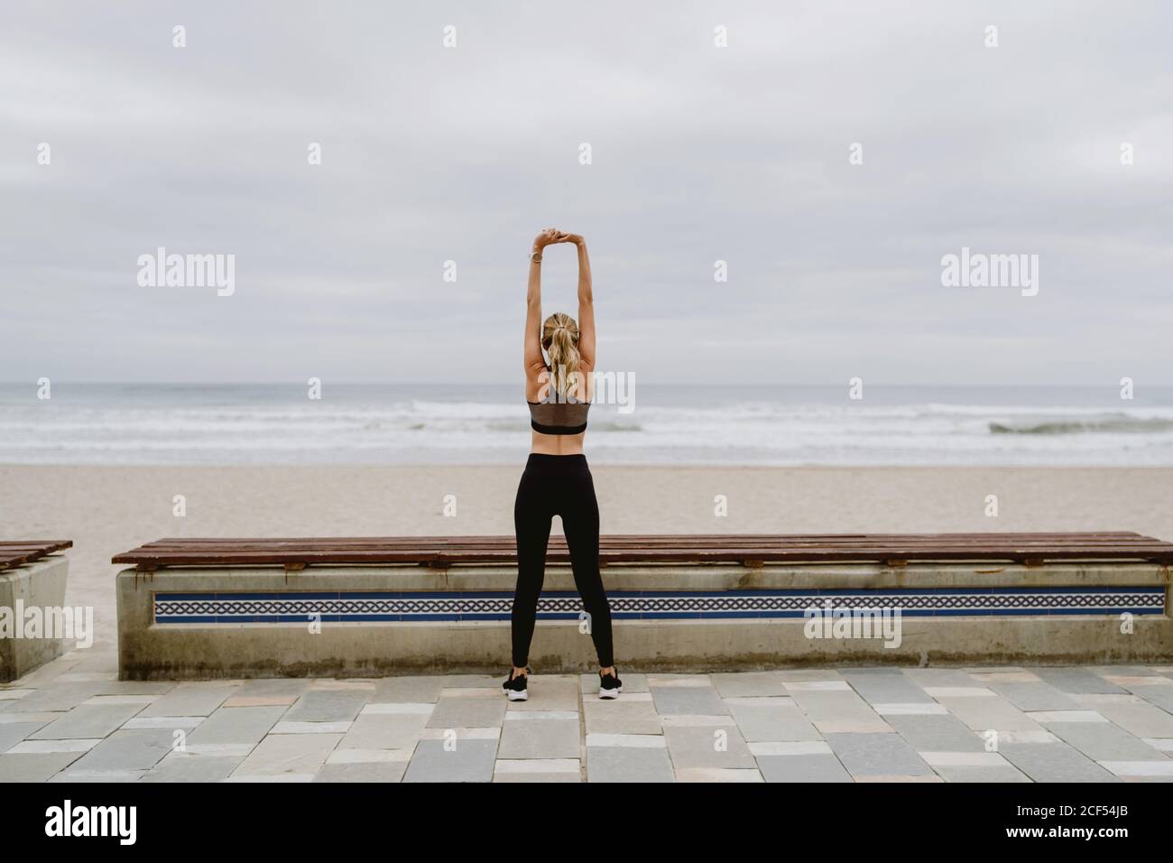Vista posteriore di atleta anonima in piedi abbigliamento attivo con le mani sollevate in blocco e che si allungano vicino al mare in tempo nuvoloso Foto Stock