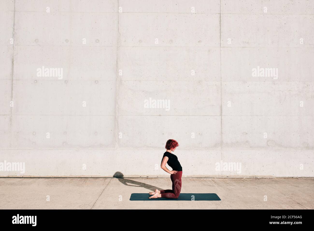 Trendy FIT Woman in sportswear fare yoga in cammello posa su materassino sportivo da solo sulla strada contro muro di cemento nella giornata di sole Foto Stock