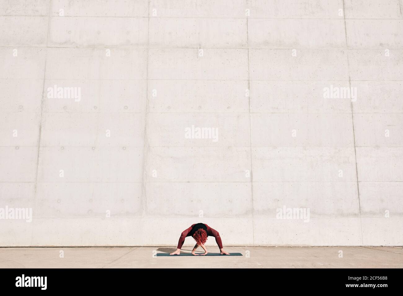 Atleta non riconoscibile in vestibilità sportiva con zampe larghe in avanti piega yoga posa su tappeto sportivo mentre allenati da solo strada contro muro di cemento in giornata di sole Foto Stock