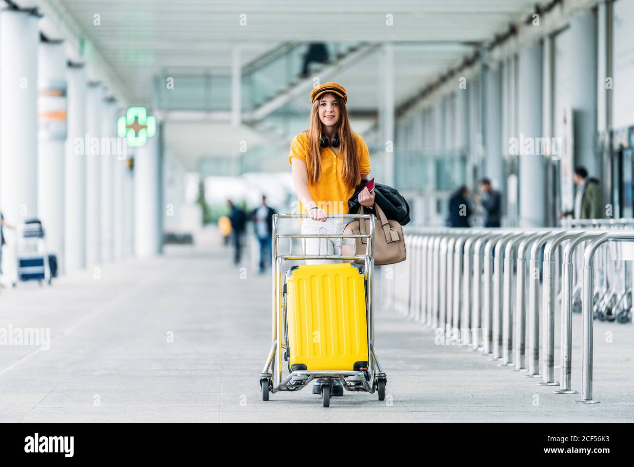 Donna sorridente in abito casual e cuffie con giacca in pelle borsa e passaporto in mano guardando la fotocamera e il trasporto cart per bagagli vicino a un moderno edificio aeroportuale Foto Stock