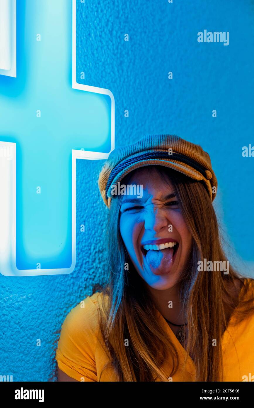 Felice giovane donna in camicia gialla e berretto alla moda fare divertente grimace e mostrare la lingua contro la parete blu con segno al neon della croce medica Foto Stock