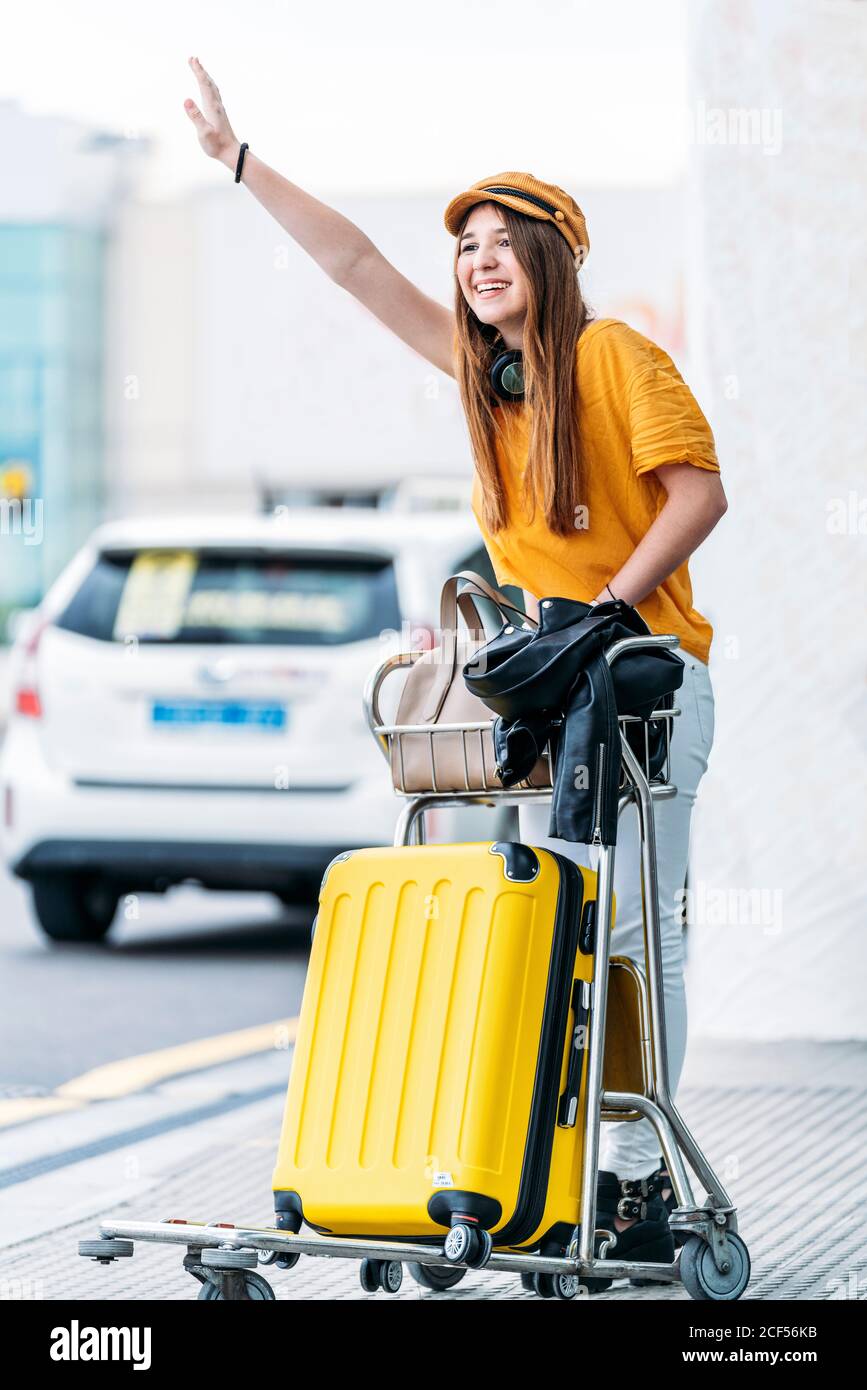 Intera lunghezza di donna allegra trendy in cappello giallo con cuffie e cart completo per il trasporto dei bagagli in taxi mentre ci si trova nelle vicinanze strada fuori dal terminal principale dell'aeroporto il caldo giorno d'estate Foto Stock