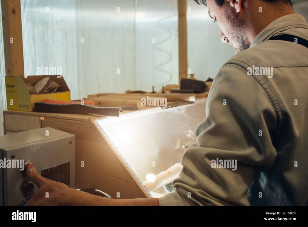 uomo in un costruttore di falegnami grigio in abiti da lavoro lavorazione di un albero con una macchina fresatrice in officina, circa un sacco di attrezzature, tavole di legno Foto Stock