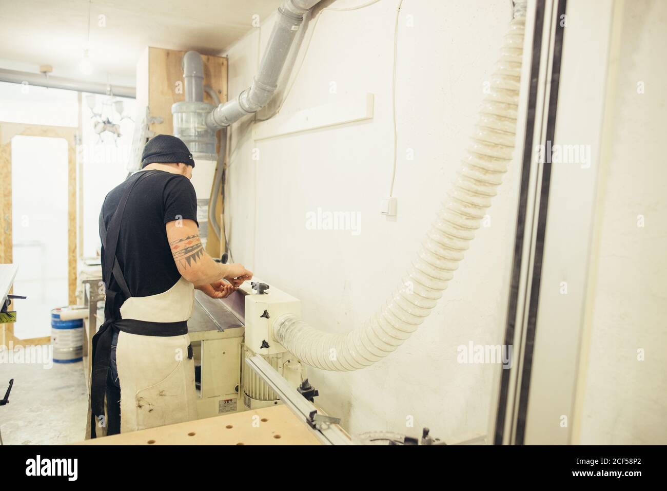 uomo in un costruttore di falegnami grigio in abiti da lavoro lavorazione di un albero con una macchina fresatrice in officina, circa un sacco di attrezzature, tavole di legno Foto Stock