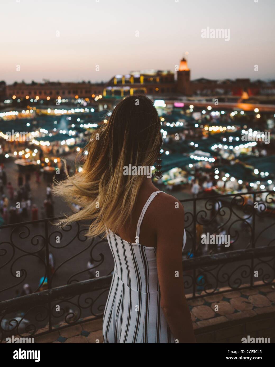 Vista posteriore della donna alla moda con i capelli volanti in piedi sul balcone contro le luci luminose della città marocchina al crepuscolo Foto Stock