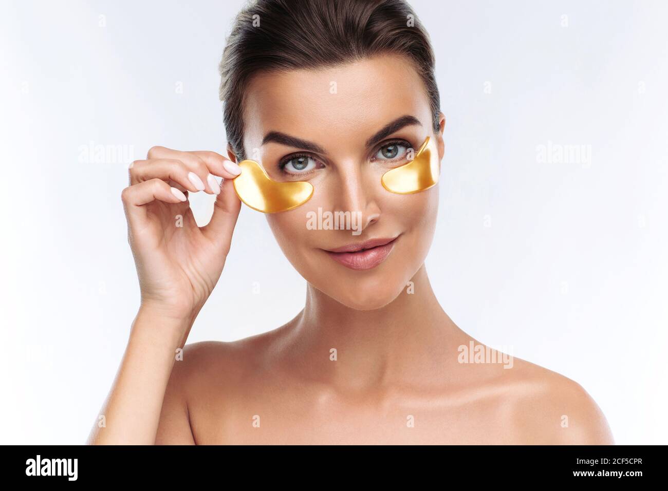 Giovane donna che applica macchie dorate di idrogel ialuronico sotto gli occhi, isolato su sfondo bianco. Cura della pelle, trattamento di bellezza per ridurre la puffiness Foto Stock