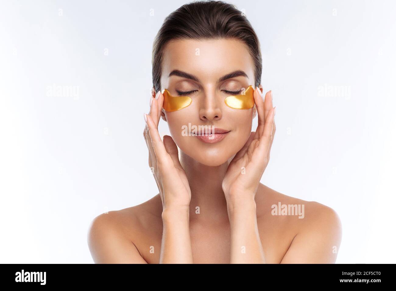 Closeup ritratto di donna di bellezza con make-up naturale applicando Golden Hydrogel cerotti sotto gli occhi. Giovane donna adulta con pelle pulita e fresca Foto Stock