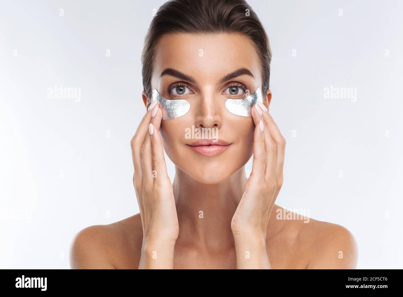 Closeup ritratto di donna di bellezza con make-up naturale applicando argentato hydrogel ialuronic cerotti sotto gli occhi. Giovane donna adulta con pelle pulita e fresca Foto Stock