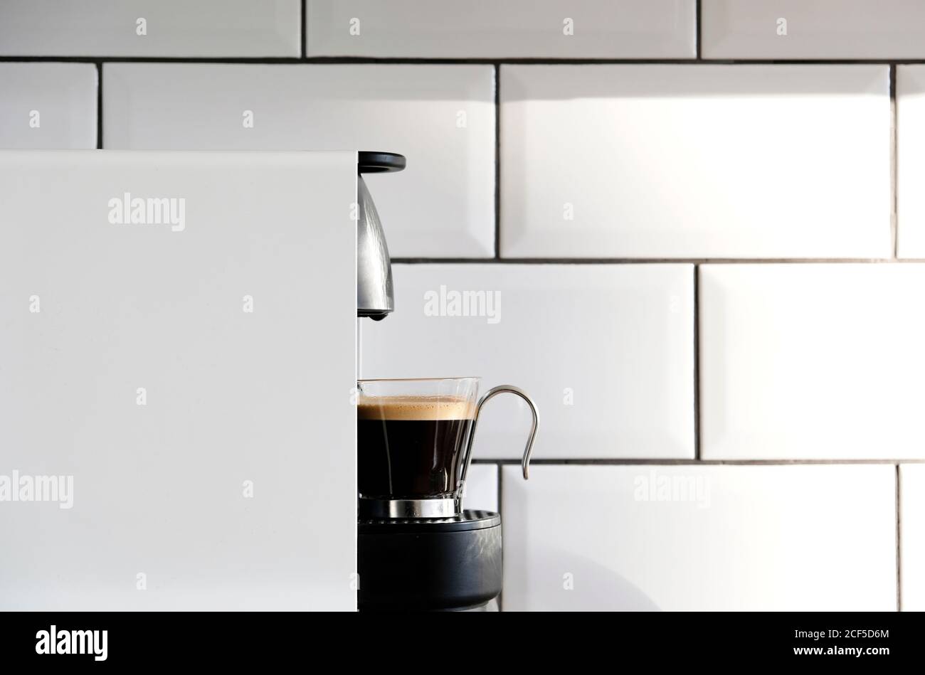 Moderna macchina per caffè a cialde che versa l'espresso caldo in una tazza di vetro su sfondo di piastrelle in ceramica su parete della cucina Foto Stock