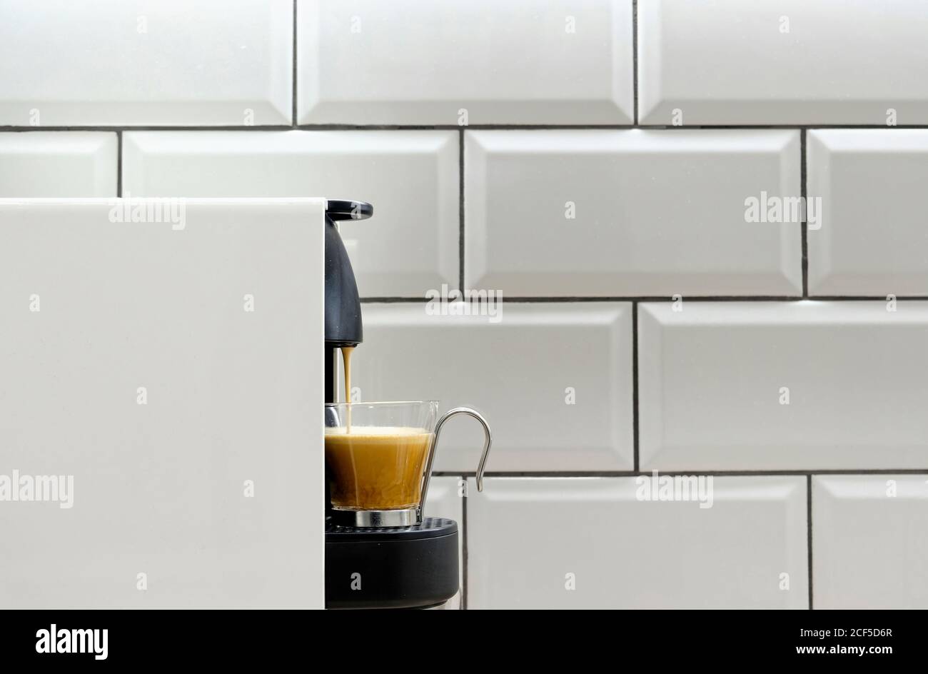 Moderna macchina per caffè a cialde che versa l'espresso caldo in una tazza di vetro su sfondo di piastrelle in ceramica su parete della cucina Foto Stock