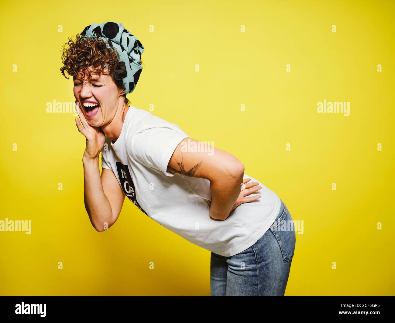 Donna adulta in abito casual alla moda con capo avvolgente occhi chiusi e ridere forte a scherzo divertente mentre in piedi su sfondo giallo Foto Stock