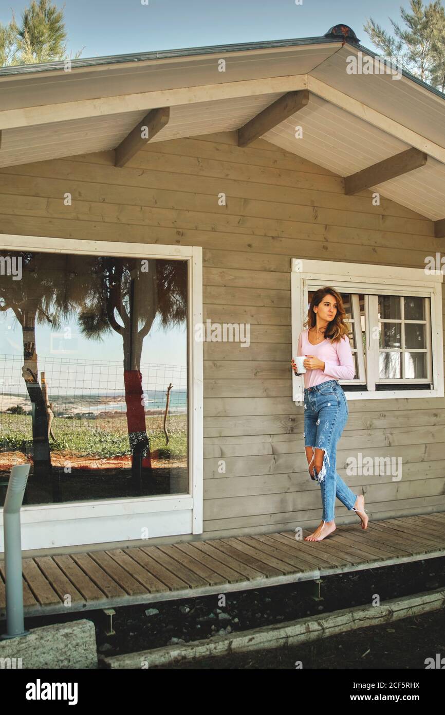 Calmo donna in jeans casual in piedi su veranda di legno di cabina accogliente e gustando una bevanda calda mentre si rilassa e si guarda via Foto Stock