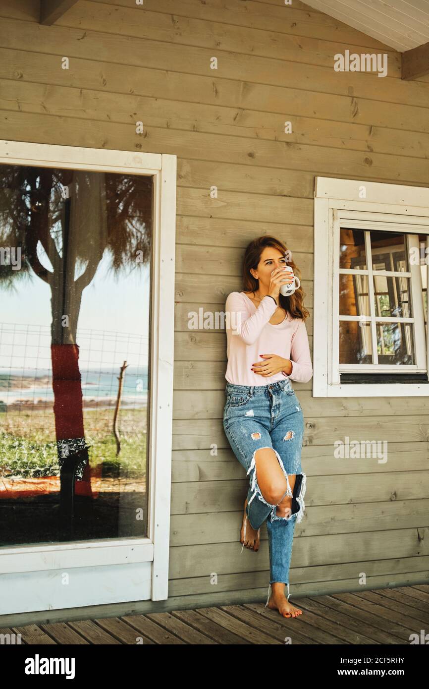 Calmo donna in jeans casual in piedi su veranda di legno di cabina accogliente e gustando una bevanda calda mentre si rilassa e si guarda via Foto Stock