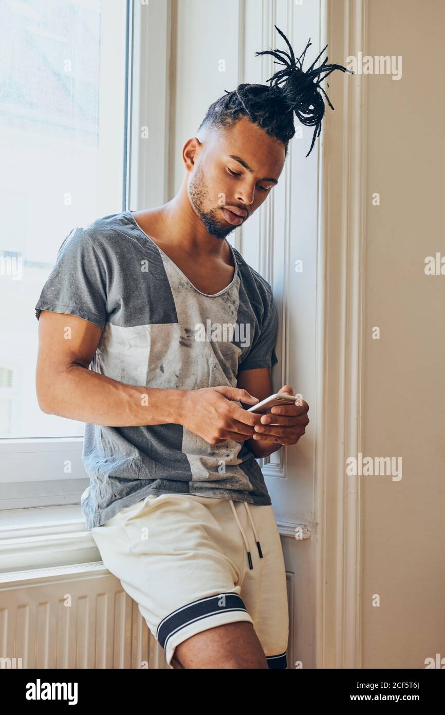 Giovane afroamericano con un'acconciatura creativa in piedi appoggiata windowsill e navigazione sul telefono cellulare Foto Stock