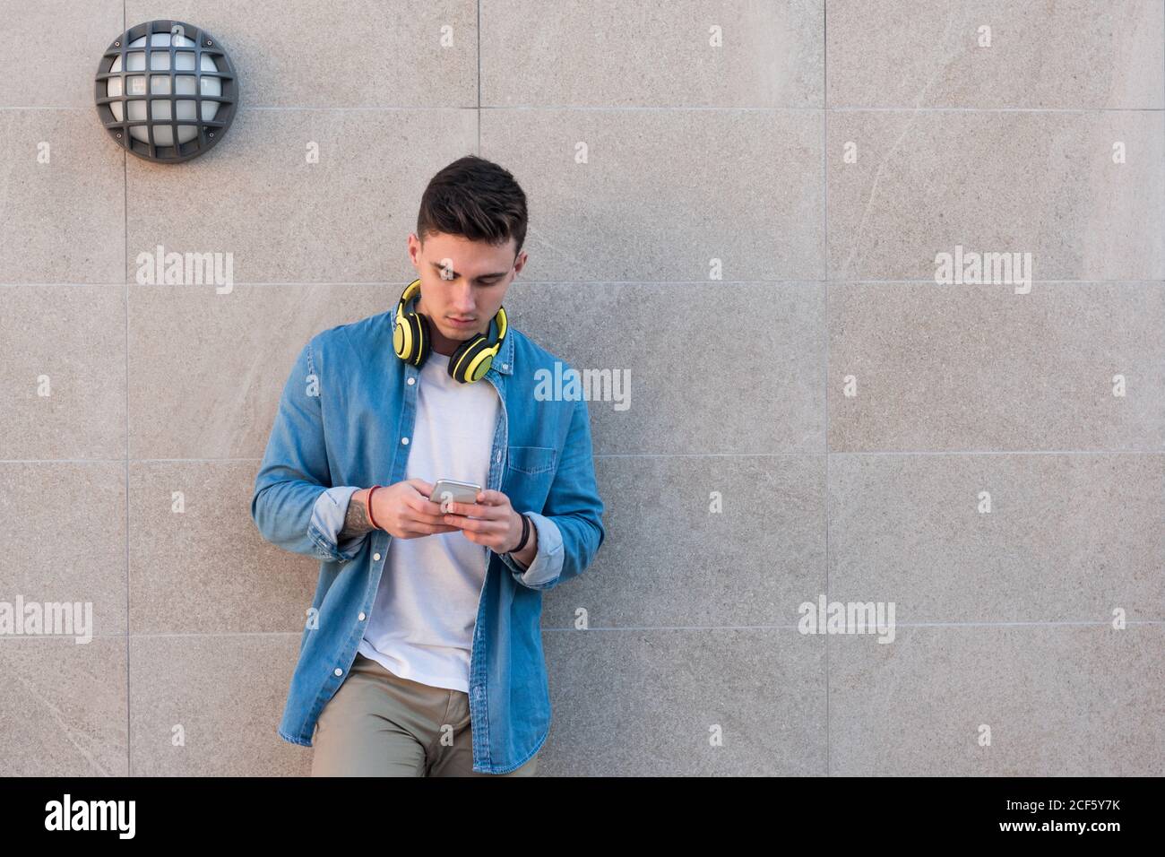 Uomo elegante e intelligente con cuffie brillanti che navigano sul telefono cellulare mentre appoggiandosi su un muro di marmo in una giornata di sole Foto Stock