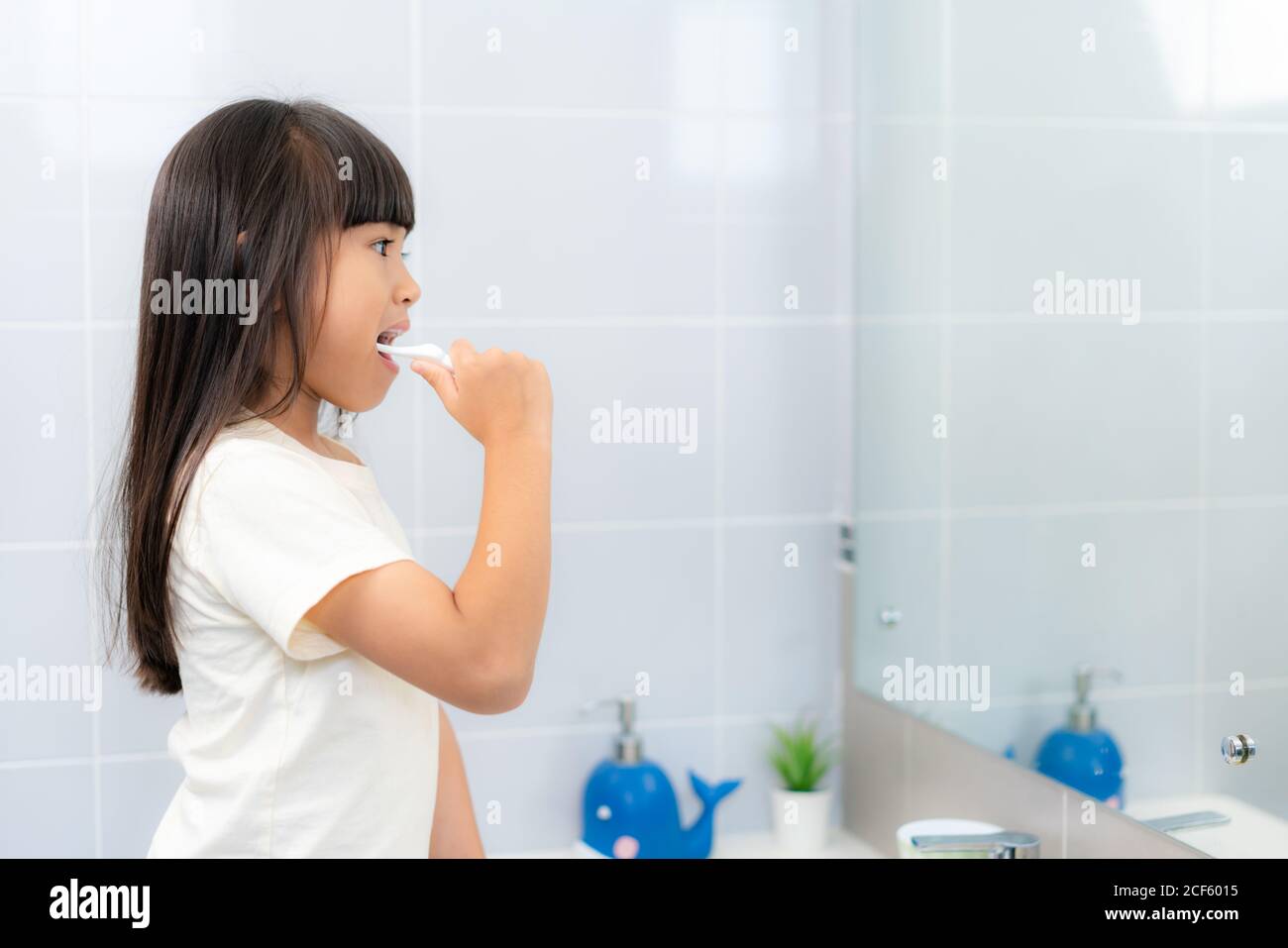 Carino asiatico scuola primaria ragazza spazzolando i denti e guardando nello specchio in bagno a casa. La routine scolastica mattutina per il giorno nella vita che si prepara Foto Stock