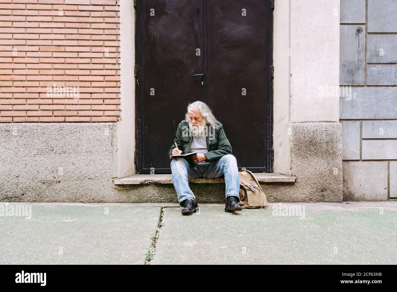 Premuroso turista maschile senior in abbigliamento casual seduto con zaino su un passo di pietra di costruzione shabby e di scrivere pensieri circa vacanza in notebook Foto Stock