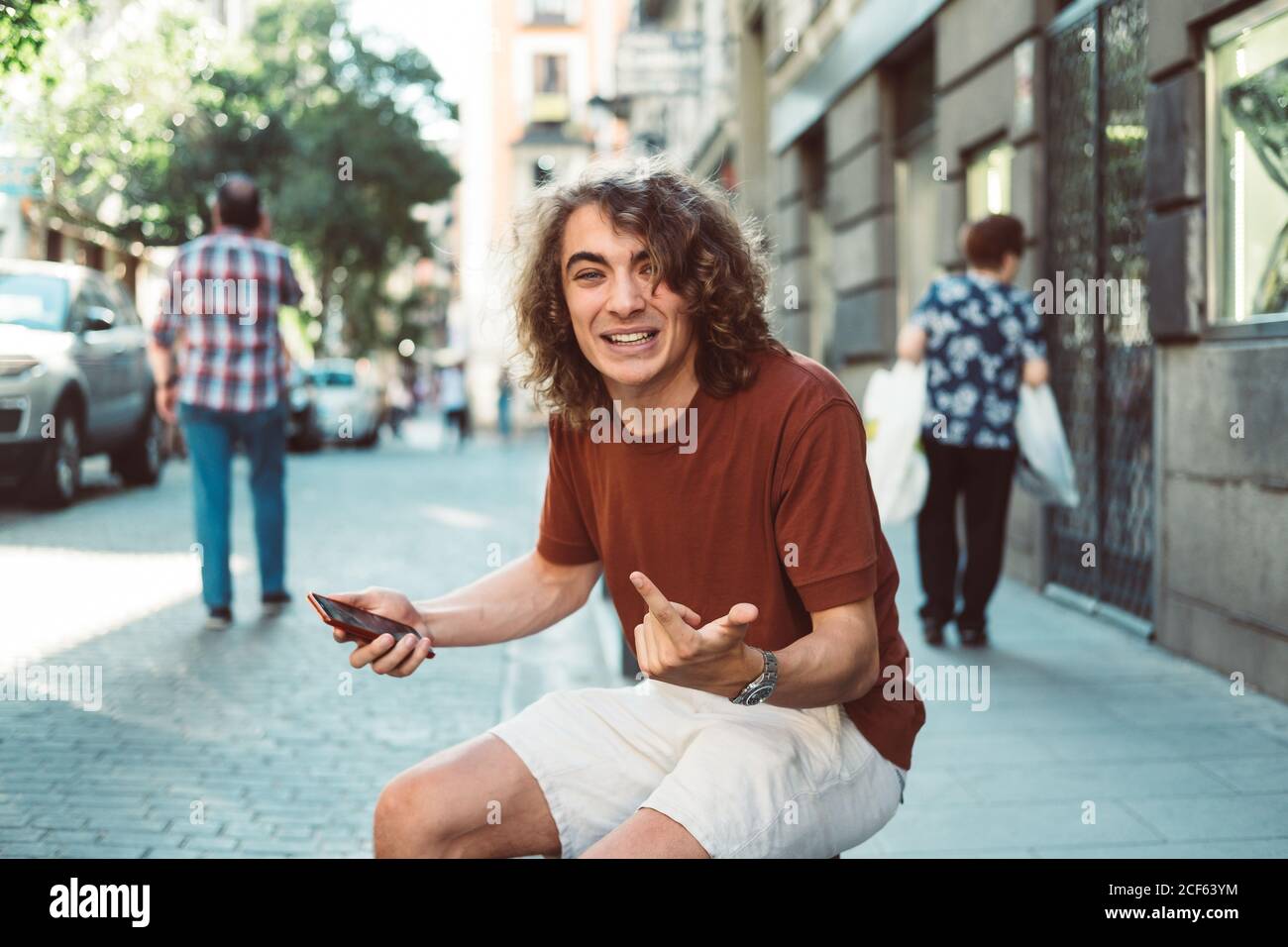 Un uomo allegro e casual ha sorpreso la creazione di volti mentre si è sullo smartphone seduto sul baluster sulla strada della città Foto Stock