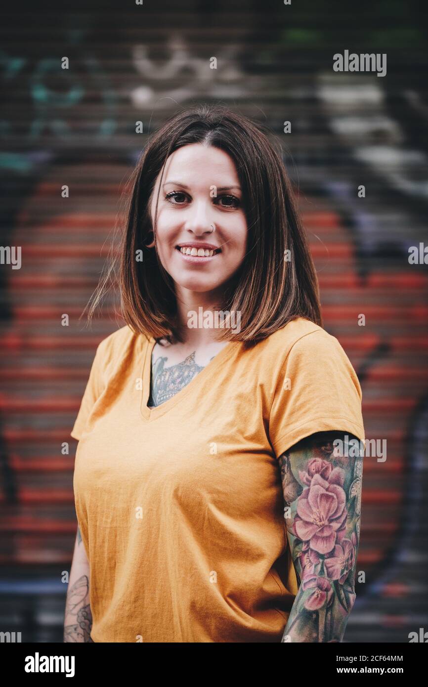 Donna moderna positiva in t-shirt sorridente mentre si trova accanto a un edificio urbano sulla strada cittadina Foto Stock