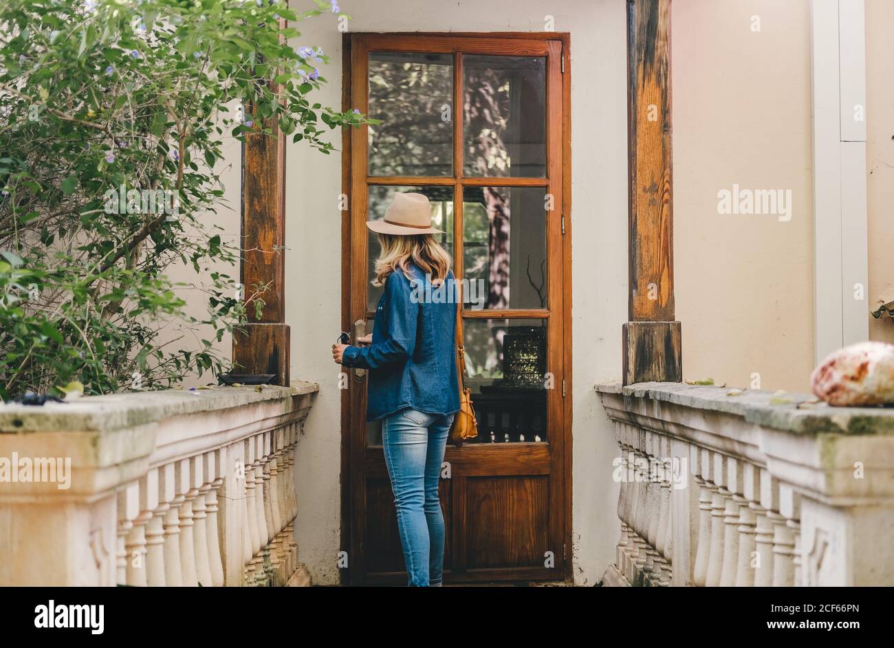 Vista posteriore della giovane donna in denim e cappello cercando di aprire la porta in vetro di legno di un edificio autentico Foto Stock