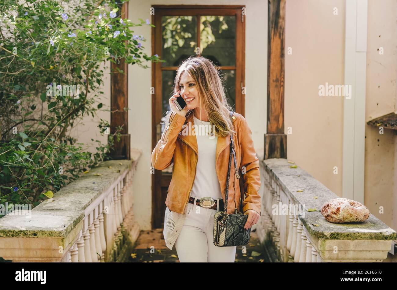 Donna moderna alla moda in giacca e jeans leggero che parla sul telefono cellulare mentre cammina sul portico di vecchia casa Foto Stock
