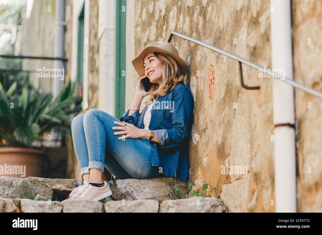 Vista laterale della giovane donna sorridente che parla sul telefono cellulare seduto su scale di pietra all'esterno Foto Stock