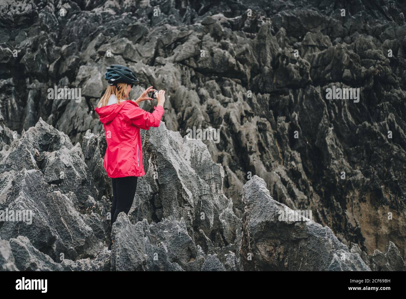 Vista laterale di Donna in casco protettivo in piedi su scogliera rocciosa e fare foto da smartphone Foto Stock