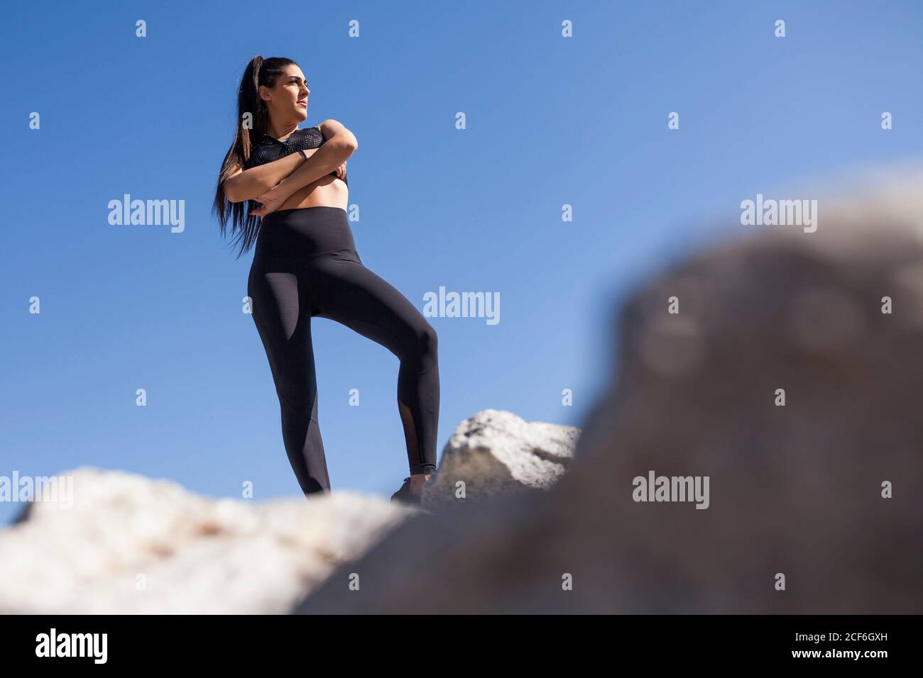 Dal basso giovane brunetta Donna in abbigliamento sportivo con mani incrociate che si posano sulle rocce nelle giornate di sole Foto Stock