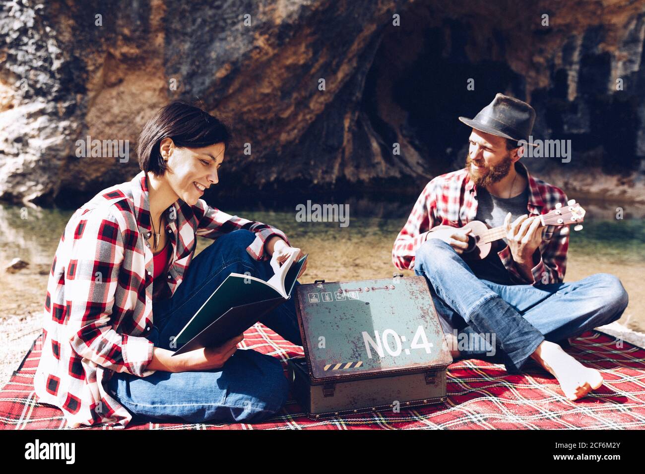 Adulto uomo e donna in plaid a scacchi con libro e piccolo ukulele pic-nic sulla riva del lago Foto Stock