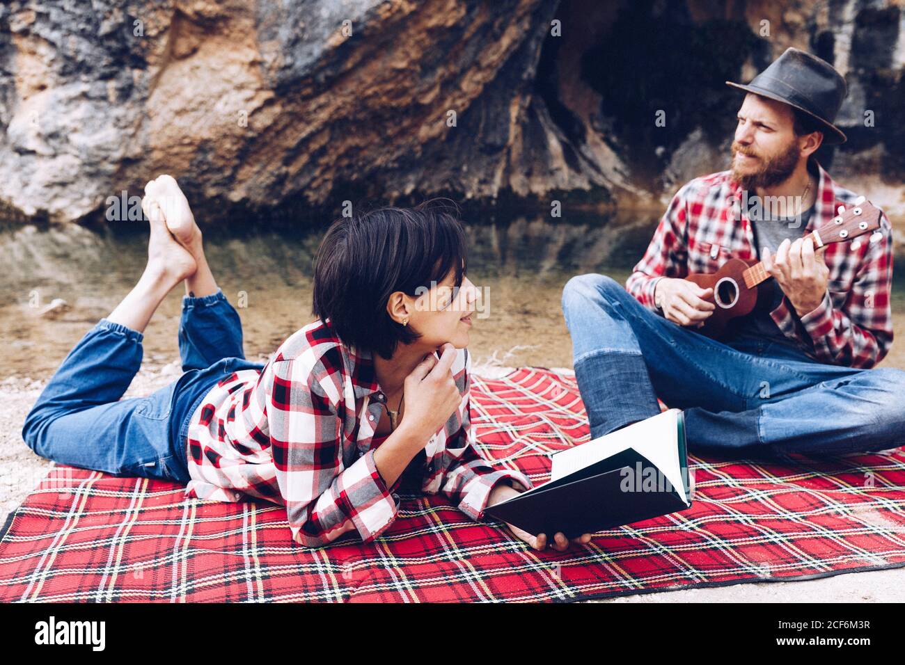 Adulto uomo e donna in plaid a scacchi con libro e piccolo ukulele pic-nic sulla riva del lago Foto Stock