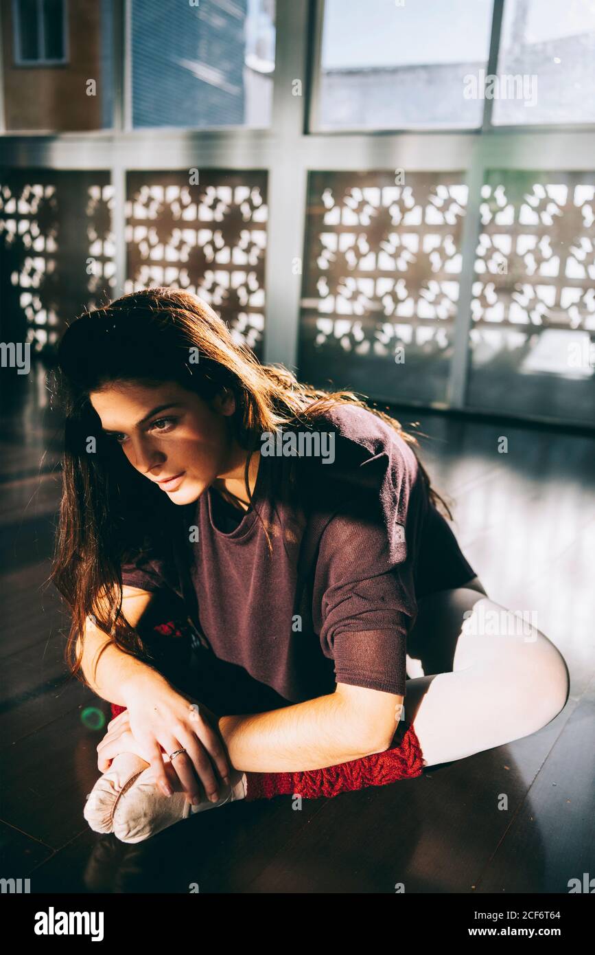 Giovane bella donna che si piega in avanti allungando le gambe mentre si siede in studio alla luce del sole. Foto Stock