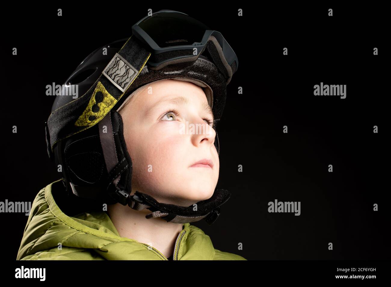 Piccolo capretto sportivo messo a fuoco in vestiti invernali con goggle di sci su sfondo nero in studio che guarda via Foto Stock