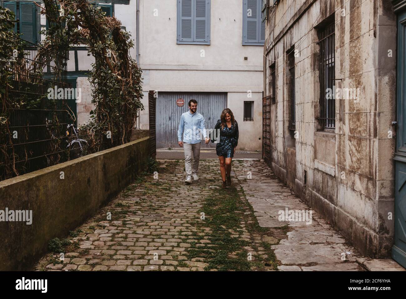 Giovane coppia allegra in abiti casual di stile che tengono le mani e. sorridente mentre si cammina lungo la vecchia strada stretta della città Foto Stock