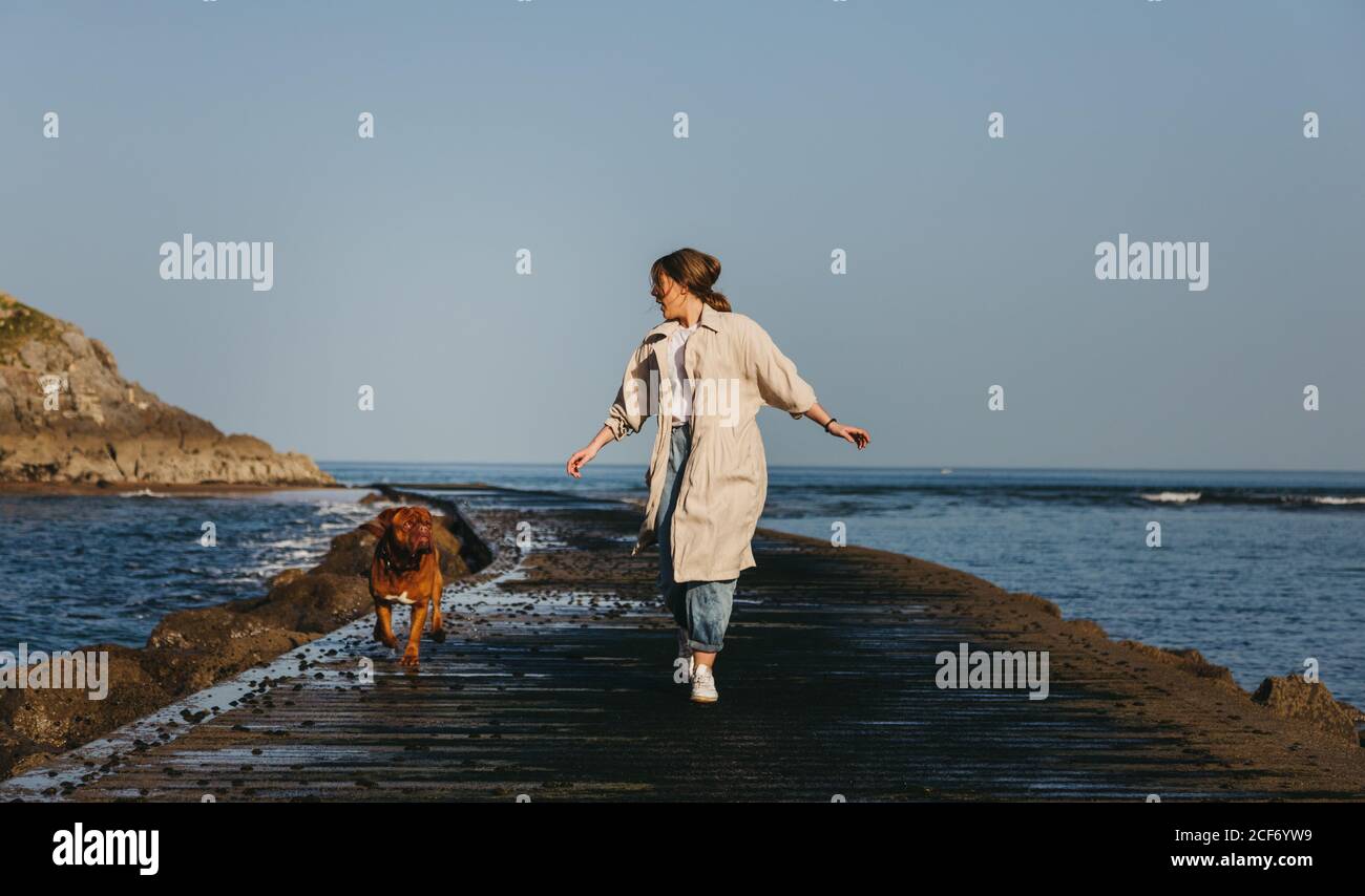 Donna in abiti casual e grande marrone Mastiff cane cercando mentre camminando lungo il molo di legno bagnato contro Calmo acqua della baia sotto il cielo blu in Spagna Foto Stock