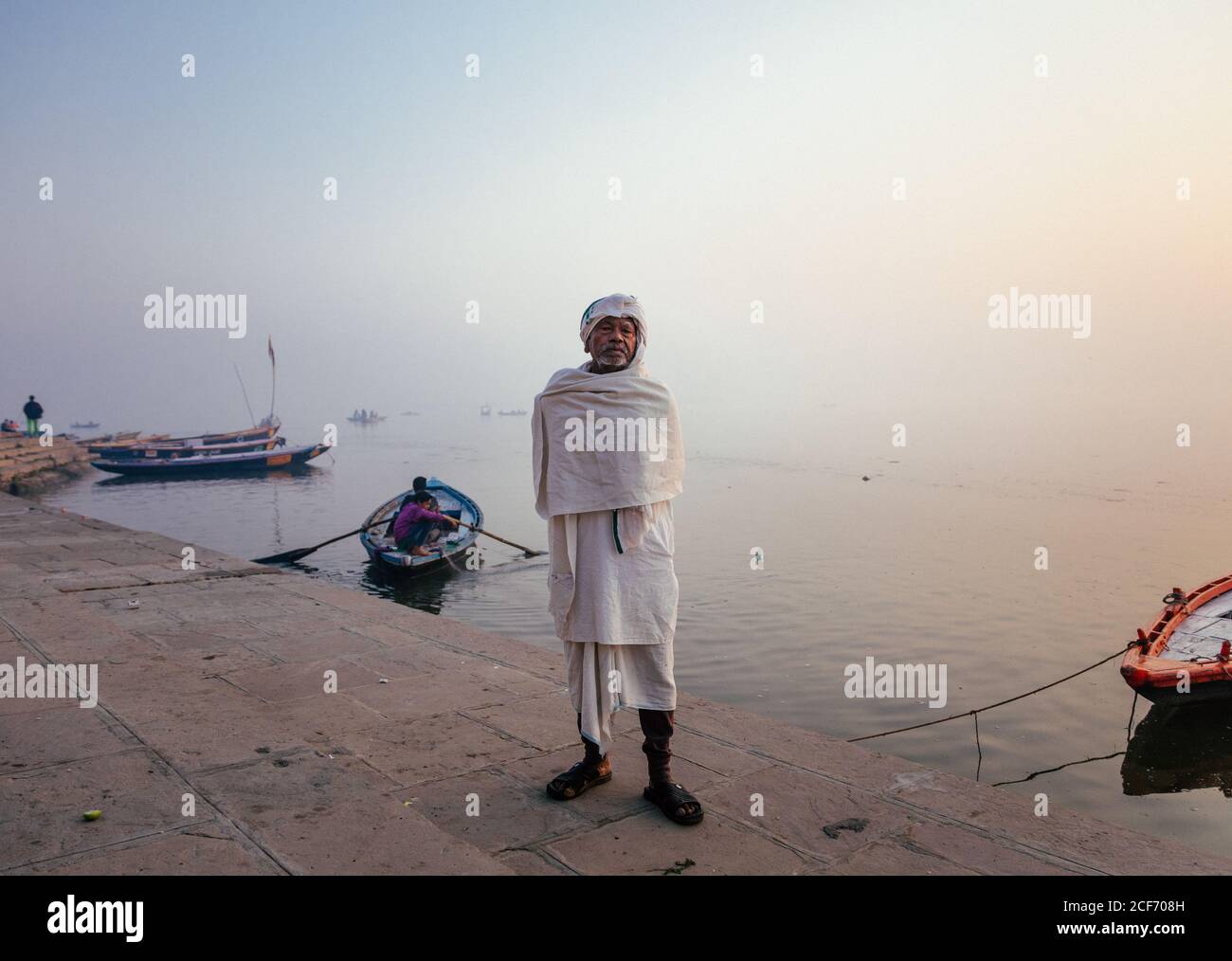 Varanasi, India - FEBBRAIO, 2018: Anziano uomo indiano che indossa abiti bianchi tradizionali in piedi sulla riva del fiume con barche da pesca in acqua nella mattina presto foggy Foto Stock