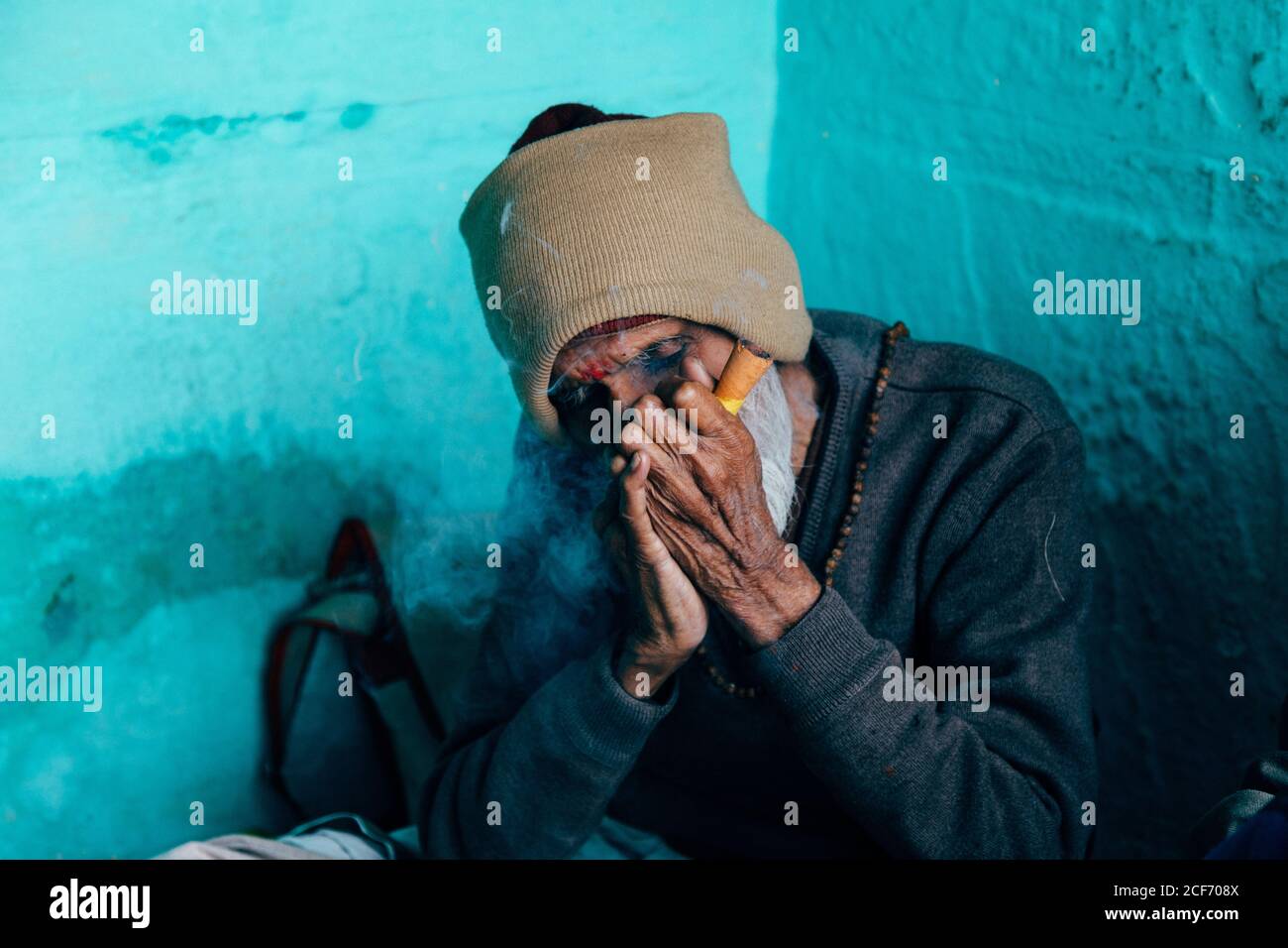 Varanasi, India - Febbraio, 2018: Povero anziano uomo indù in berretto lavorato beige seduto nell'angolo della casa con pareti blu e sigaro fumante Foto Stock