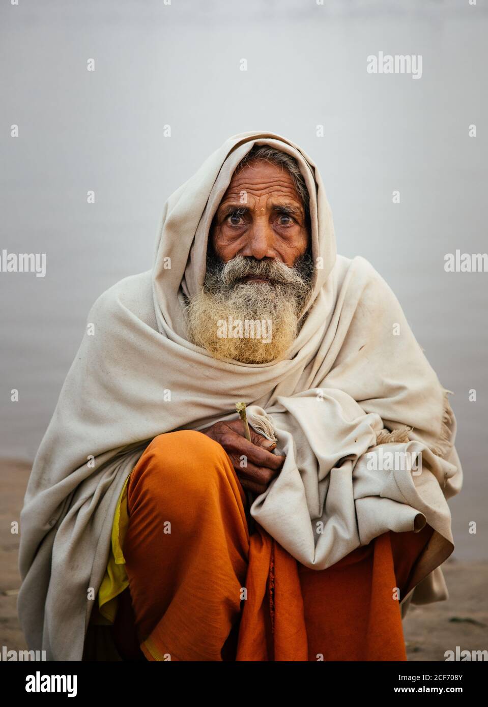 Varanasi, India - FEBBRAIO, 2018: Anziano uomo indiano che indossa abiti tradizionali arancioni seduti sulla riva del fiume con barche da pesca in acqua nella mattina presto foggy Foto Stock