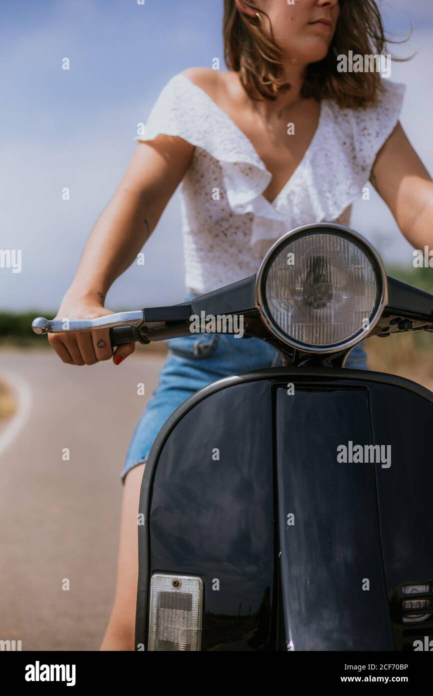 Crop giovane Donna in abiti casual guida scooter su strada il giorno di sole Foto Stock