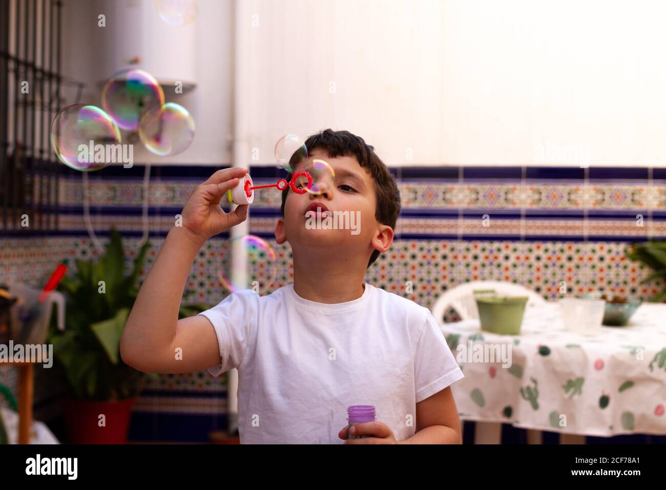 Bambino in T-shirt bianca che soffia bolle di sapone mentre si è in piedi sulla terrazza a casa Foto Stock