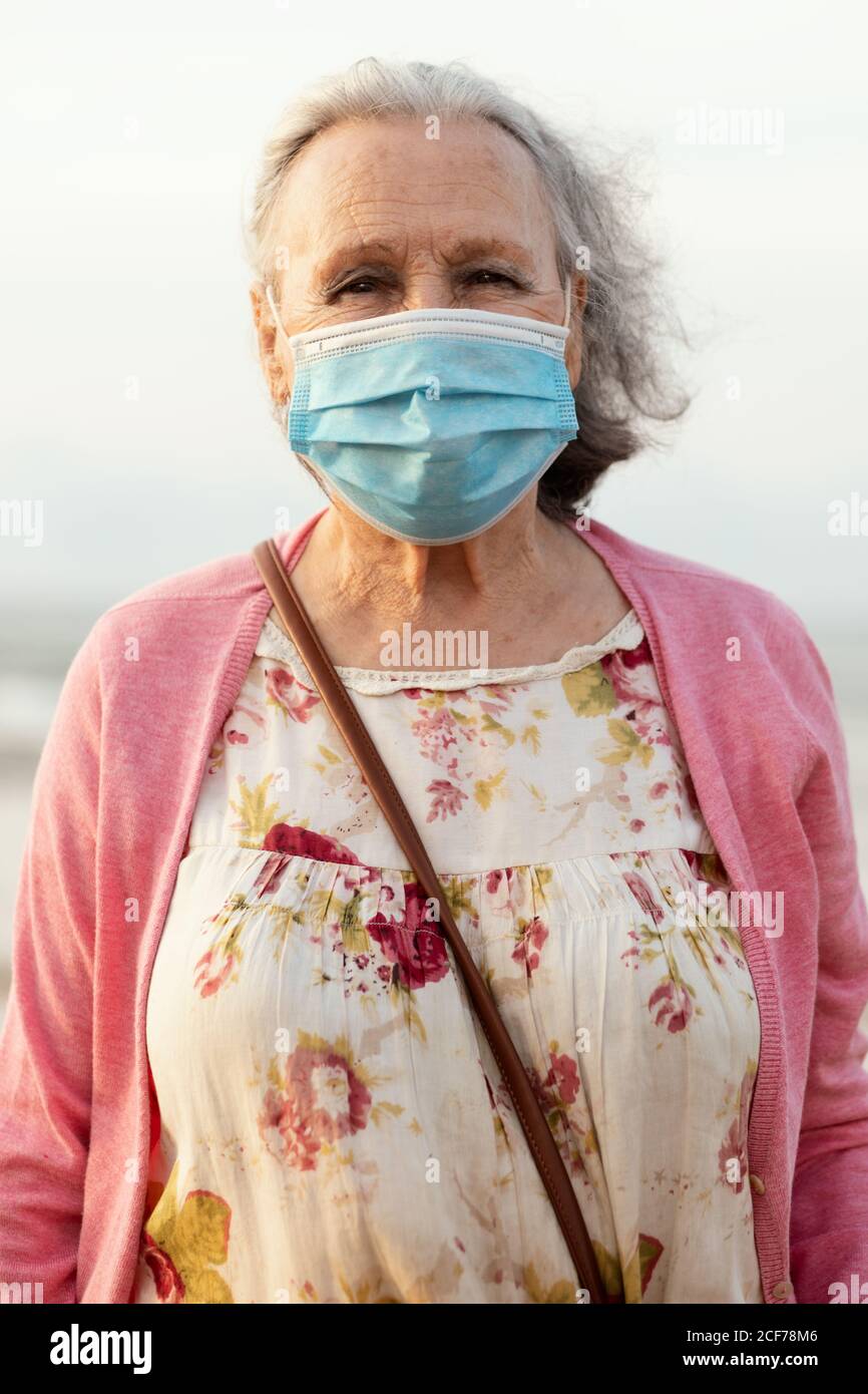 Donna senior con capelli grigi in maschera blu sterile e casual vestiti guardando la fotocamera mentre si è in piedi su sfondo sfocato in luce del giorno Foto Stock