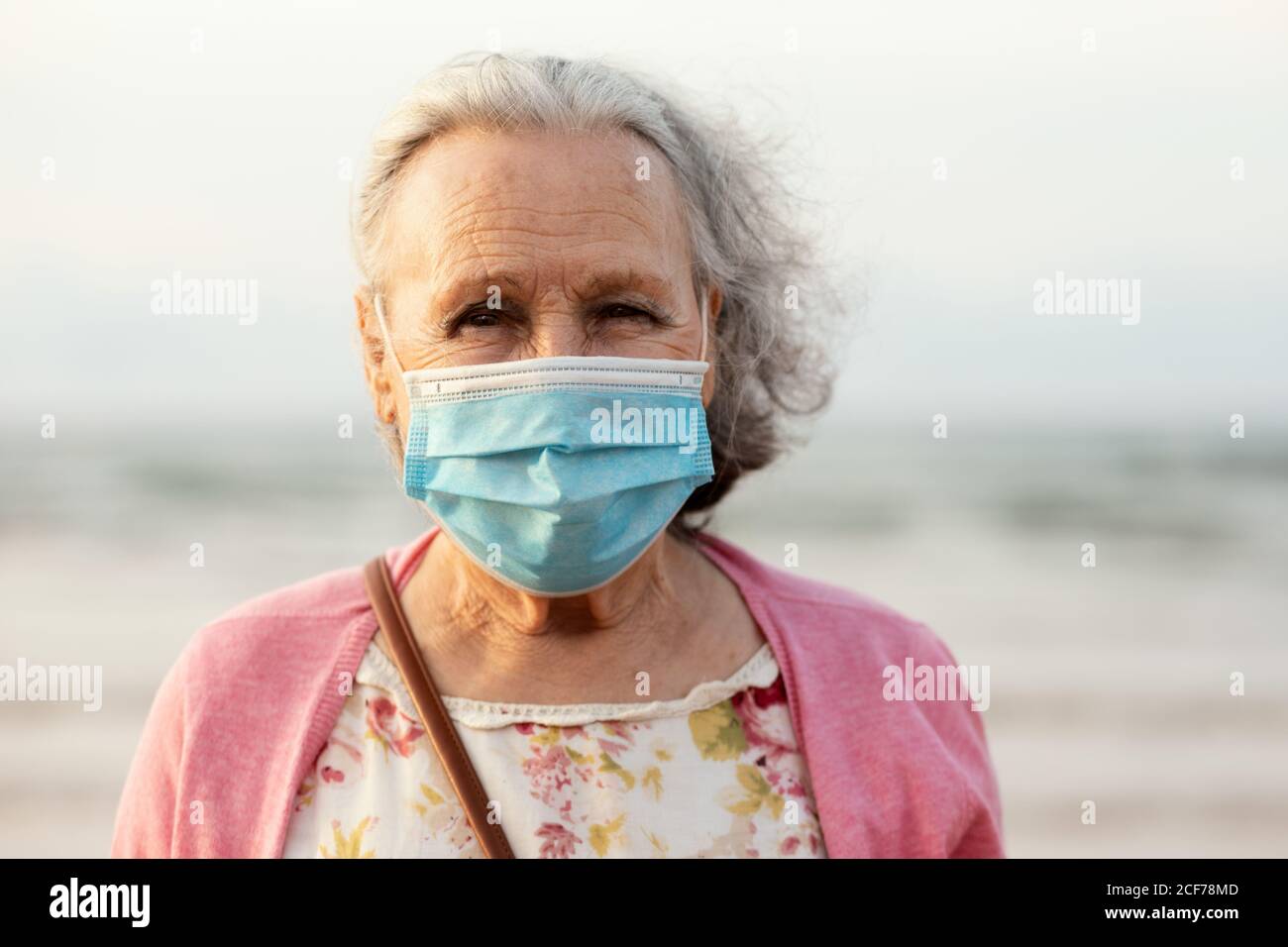 Donna senior con capelli grigi in maschera blu sterile e casual vestiti guardando la fotocamera mentre si è in piedi su sfondo sfocato in luce del giorno Foto Stock