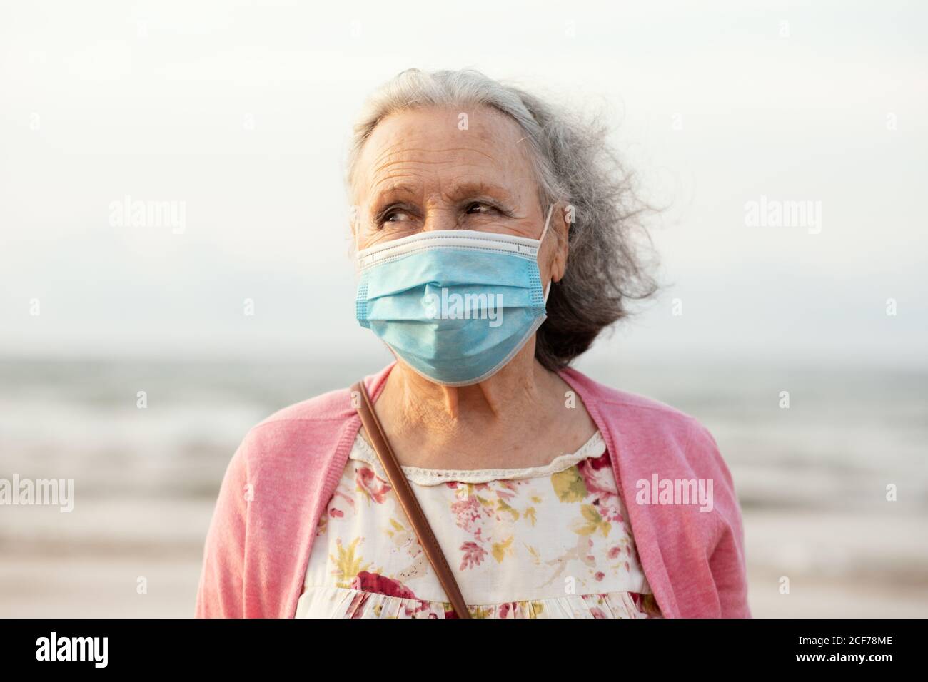 Donna senior con capelli grigi in maschera blu sterile e casual vestiti che guardano via mentre si levano in piedi su sfondo sfocato nella luce del giorno Foto Stock