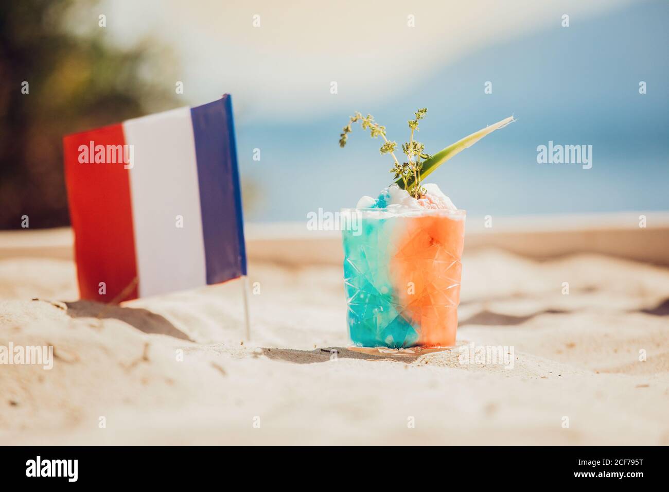 Bicchiere di bevanda alcolica rinfrescante nei colori della bandiera francese guarnito con cubetti di ghiaccio ed erbe poste sulla spiaggia di sabbia contro il mare offuscato Foto Stock