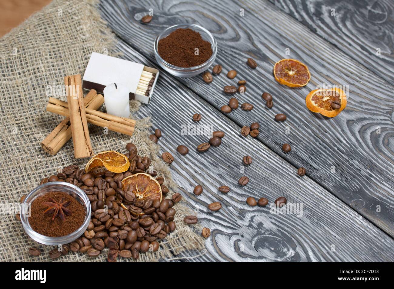 Chicchi di caffè su tela ruvida e stub di candela. Caffè macinato in contenitori e spezie. Su una superficie di tavole di pino spazzolato dipinte in bianco e nero. Foto Stock