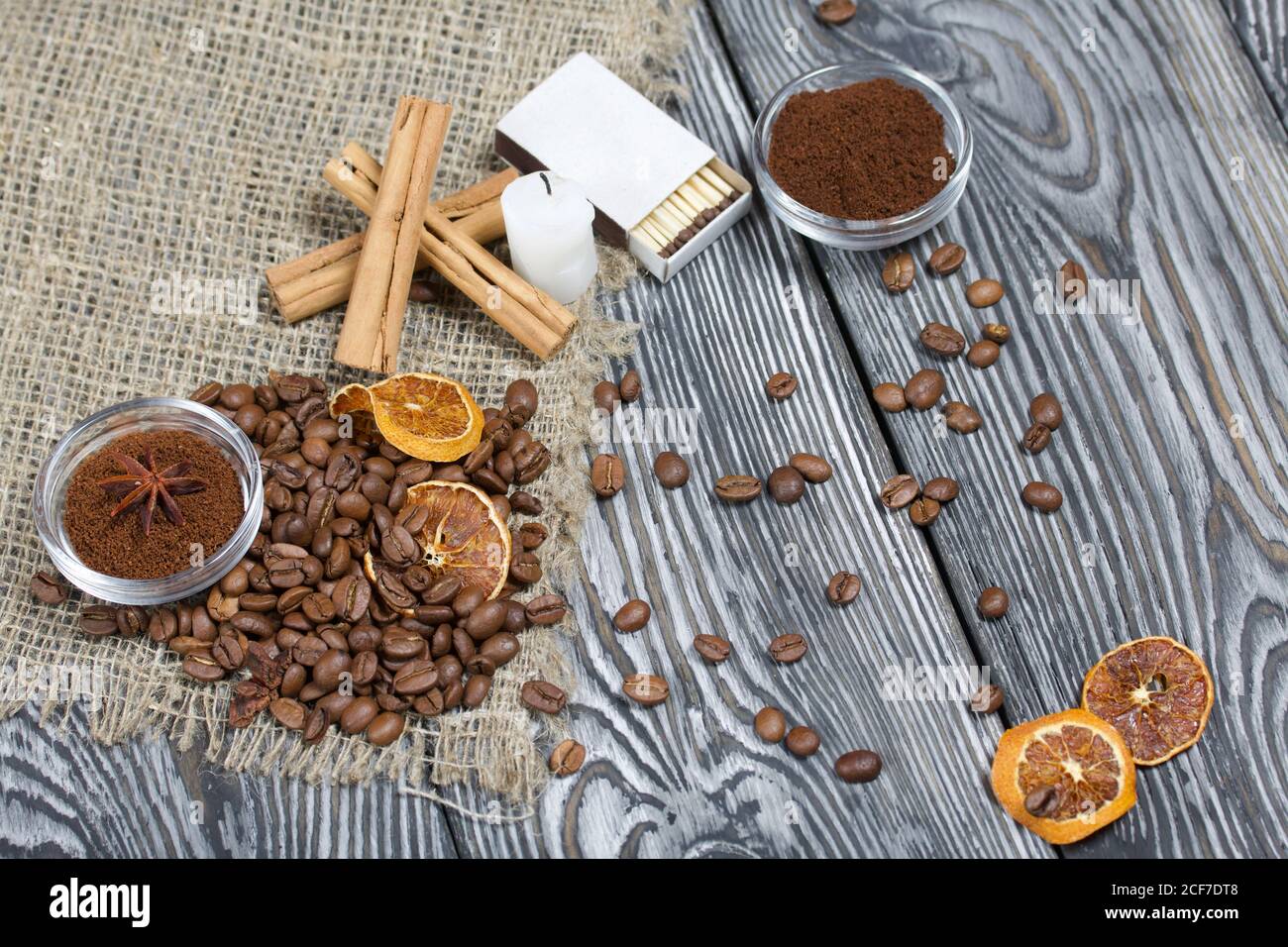 Chicchi di caffè su tela ruvida e stub di candela. Caffè macinato in un contenitore e spezie. Su una superficie di tavole di pino spazzolato dipinte in bianco e nero. Foto Stock
