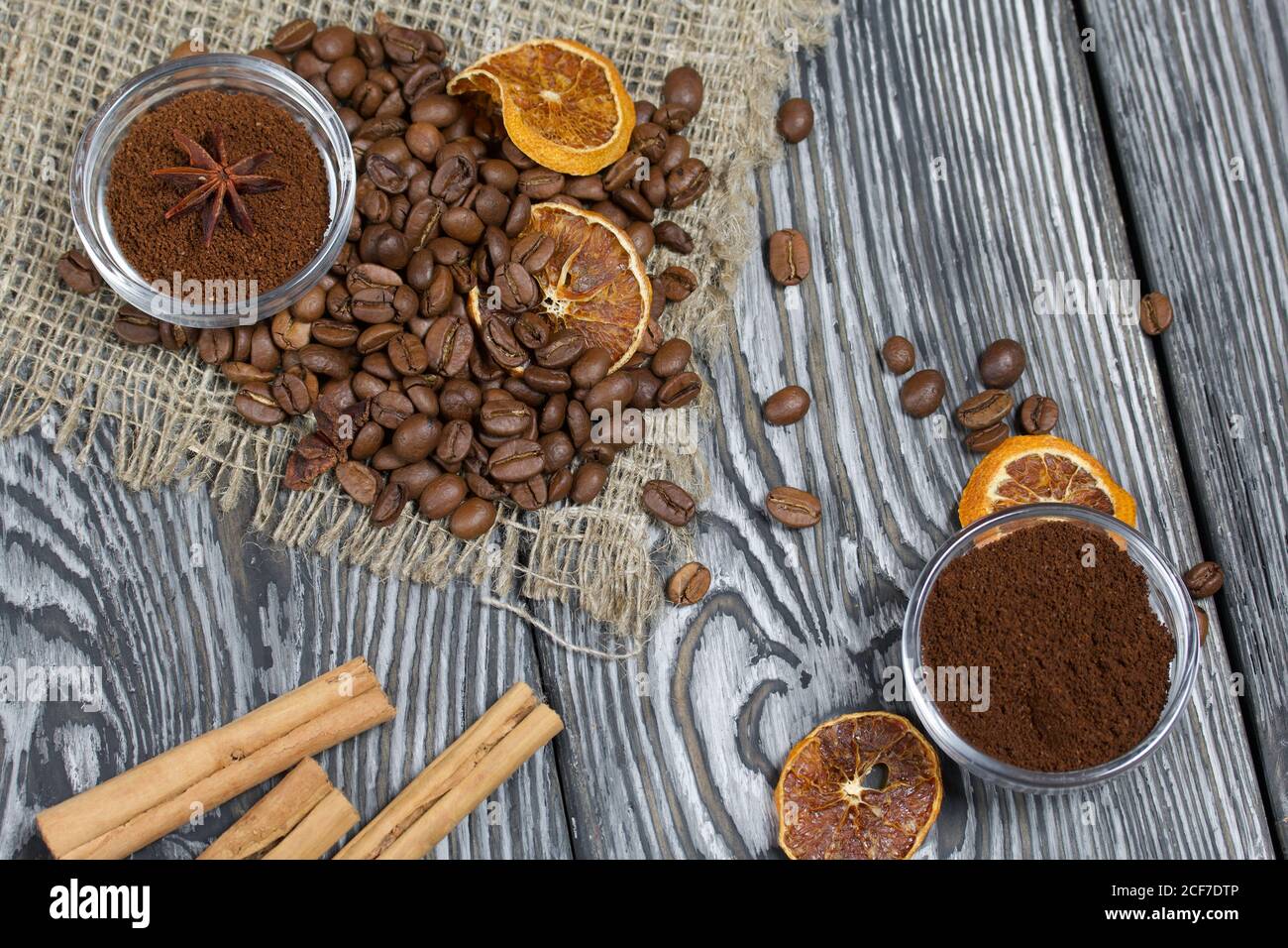Chicchi di caffè su lino grosso. Caffè macinato in contenitori e spezie. Su una superficie di tavole di pino spazzolato dipinte in bianco e nero. Foto Stock