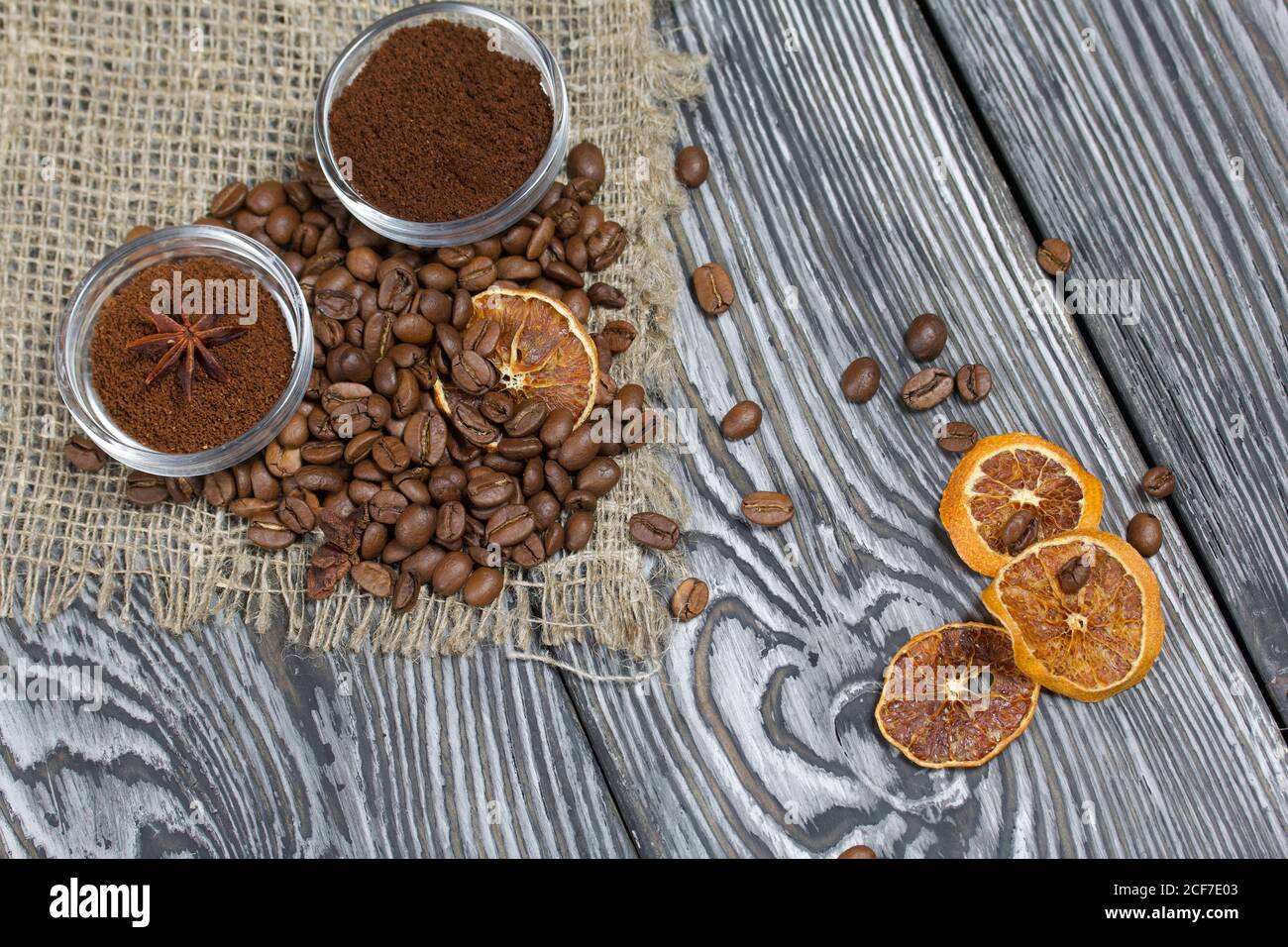 Chicchi di caffè su lino grosso. Caffè macinato in contenitori e spezie. Su una superficie di tavole di pino spazzolato dipinte in bianco e nero. Foto Stock