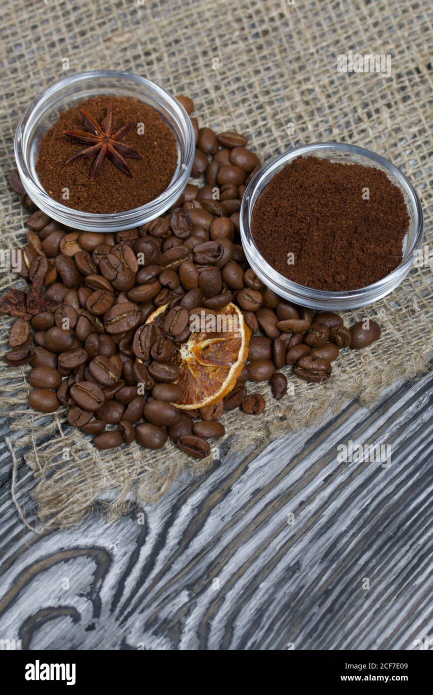 Chicchi di caffè su lino grosso. Caffè macinato in contenitori e spezie. Su una superficie di tavole di pino spazzolato dipinte in bianco e nero. Foto Stock