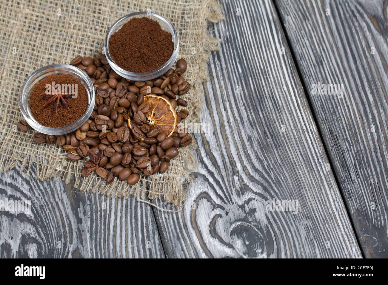 Chicchi di caffè su lino grosso. Caffè macinato in contenitori e spezie. Su una superficie di tavole di pino spazzolato dipinte in bianco e nero. Foto Stock